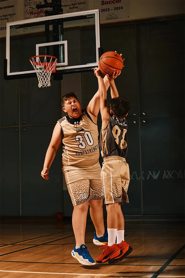 Two young basketball players in beige uniforms competing for the ball mid-air under a basketball hoop in a gym.