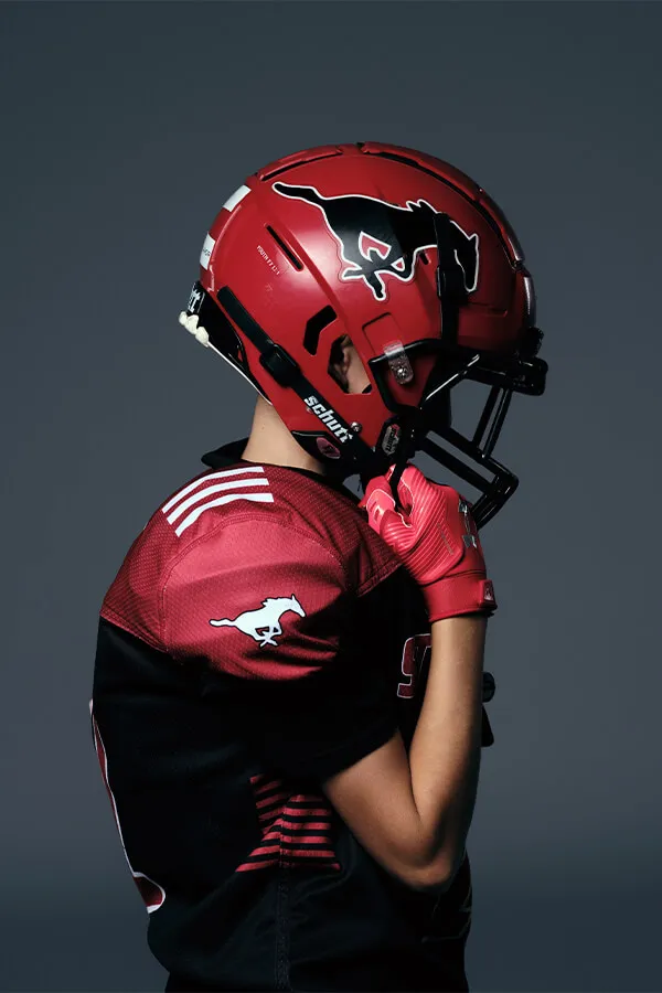 Side profile of a football player wearing a red helmet with a black mustang logo and matching red and black jersey and gloves against a dark background.