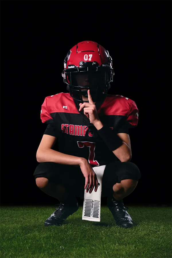 Football player in red and black uniform crouching on grass with finger to helmet visor indicating silence.