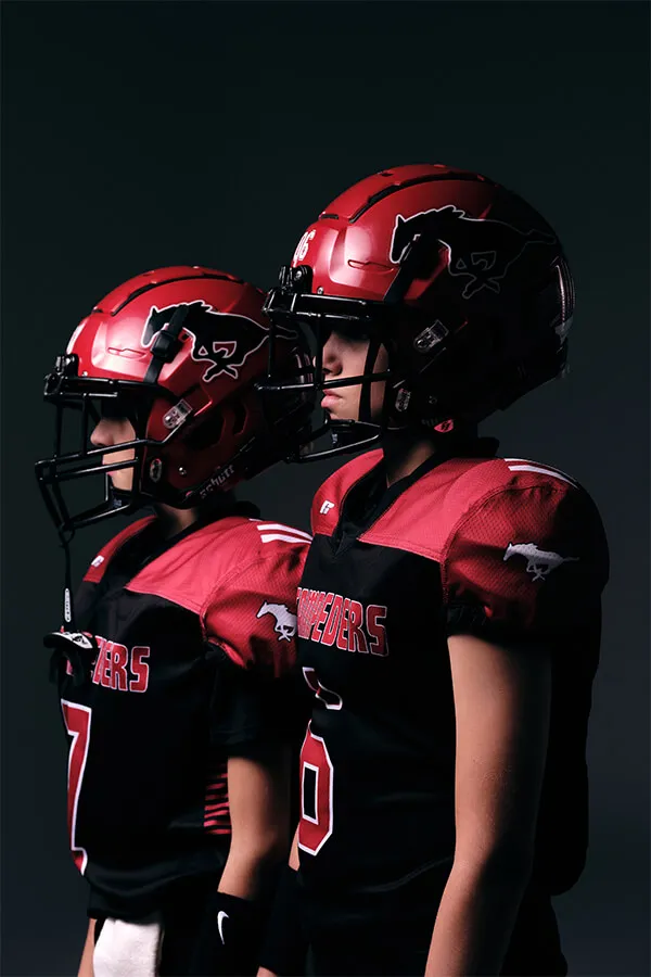 Two youth football players in red and black uniforms and helmets standing side by side against a dark background.