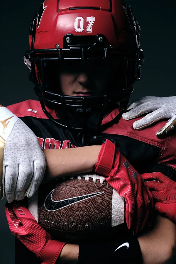 American football player wearing a red helmet marked '07' holding a Nike football, with white-gloved hands on his shoulders.
