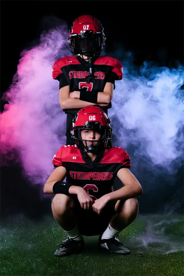 Two young football players in red and black Stampeders uniforms and helmets posing with colorful smoke in the background.