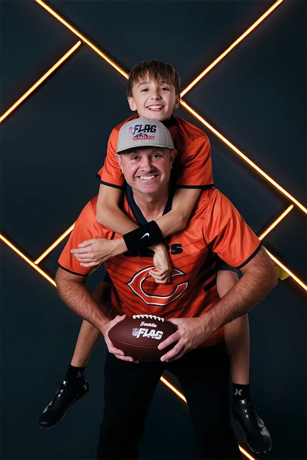 Man wearing a gray NFL FLAG cap and orange shirt holding a football with a boy in an orange shirt on his back against a dark background with orange diagonal lights.
