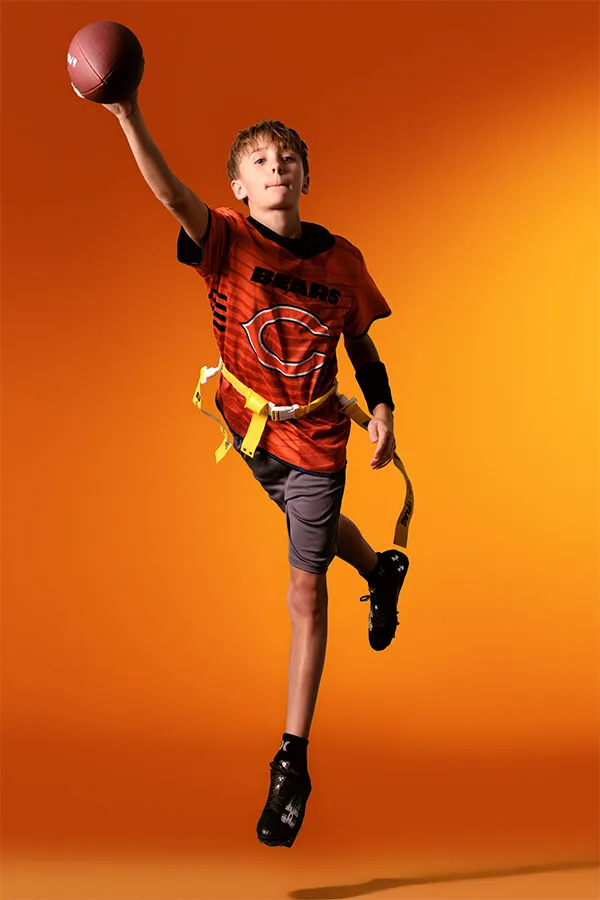 Boy in an orange Bears jersey jumping and throwing a football against an orange background.