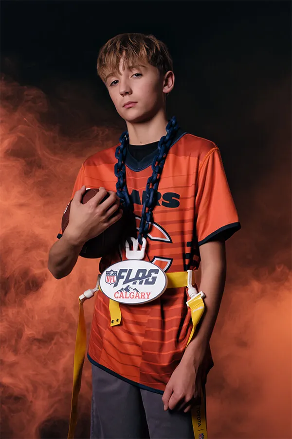 Teen boy in an orange sports jersey holding a football with an NFL Flag Calgary belt and yellow flag football flags against a smoky dark background.