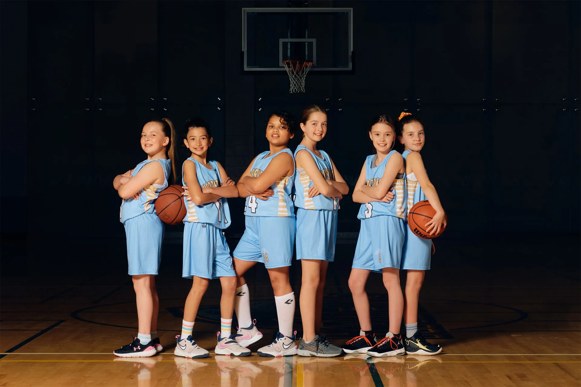 Six girls in blue basketball uniforms standing back to back on an indoor court with two holding basketballs.
