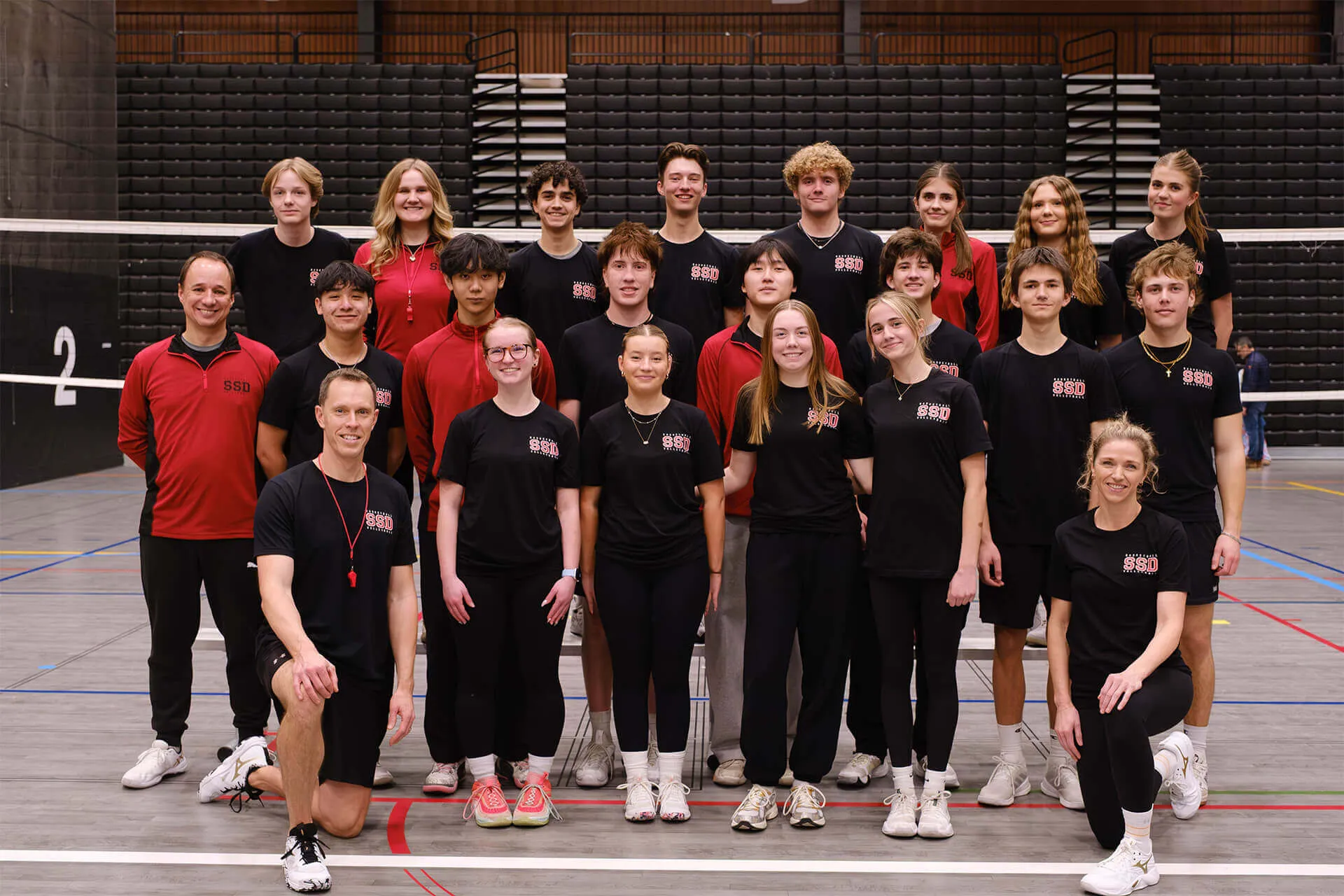 Group photo of a volleyball team and coaches posing in a gym with a net and bleachers in the background.