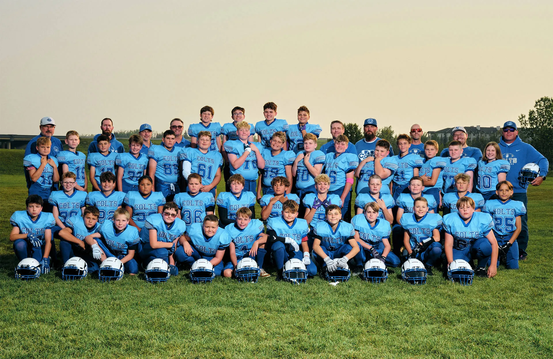 Youth football team in blue Colts uniforms posing outdoors on grass with helmets lined up in front, accompanied by their coaches.