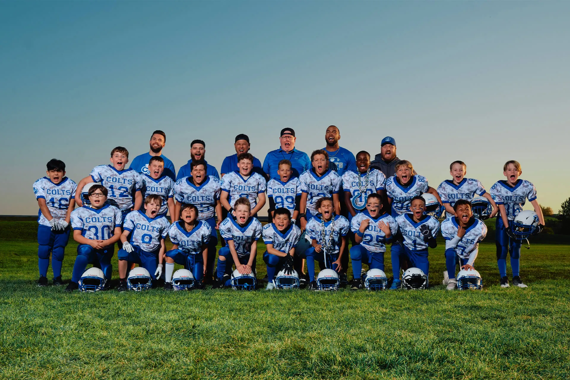 Youth football team in blue and white Colts uniforms posing enthusiastically on grass field at sunset.