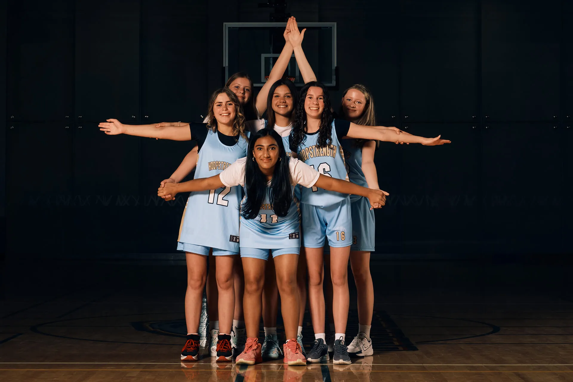 Six female basketball players in light blue uniforms posing in a basketball court with arms extended in a formation.