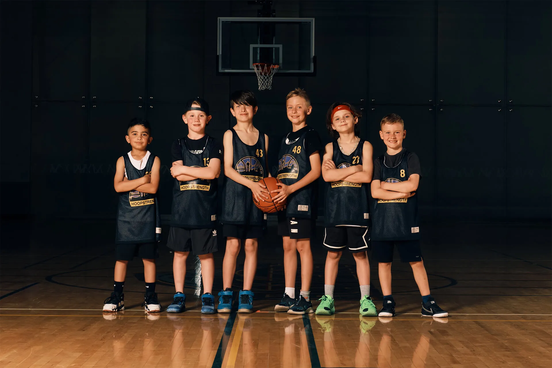 Six young boys in black basketball jerseys standing with arms crossed on an indoor court, with one boy holding a basketball.
