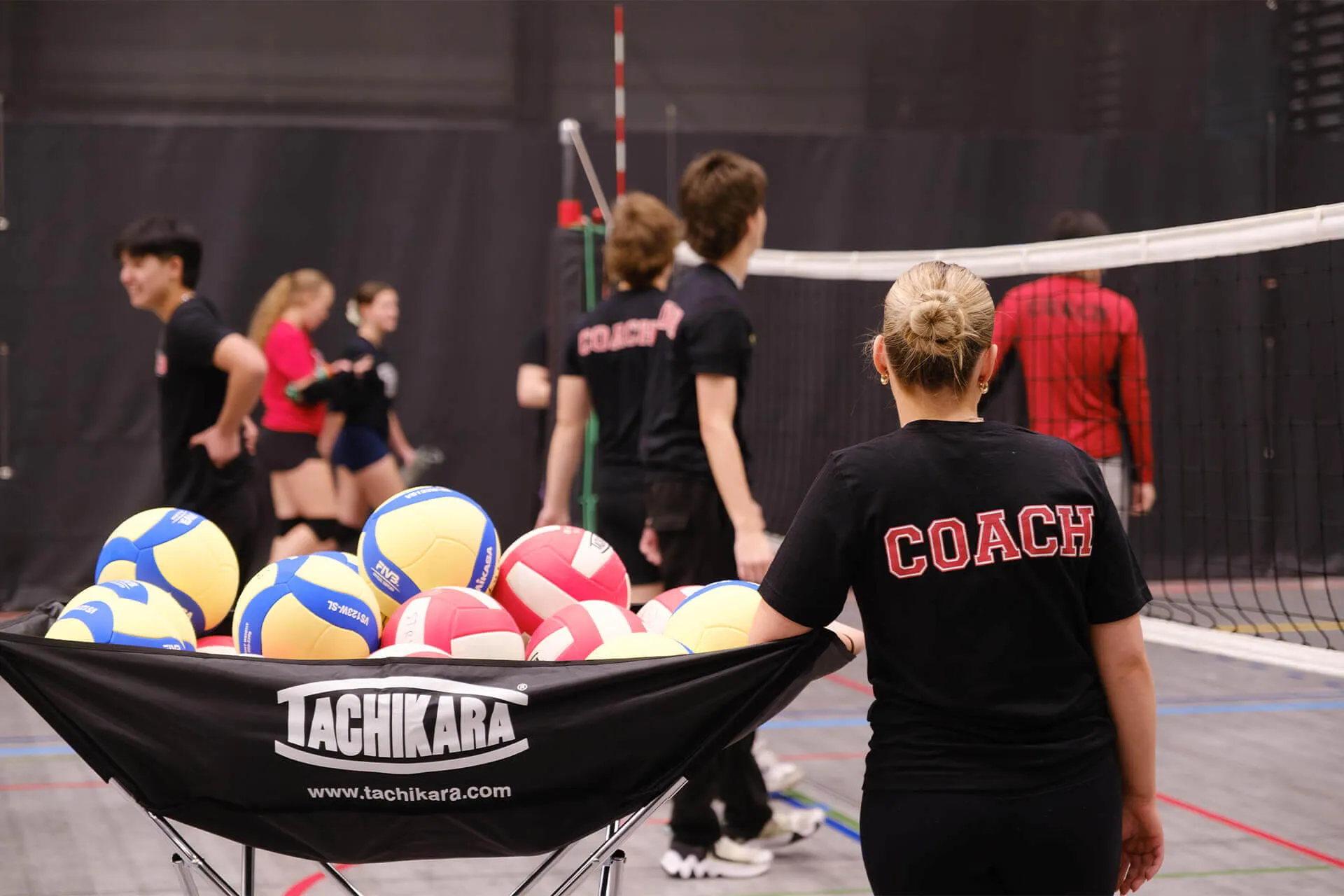 Volleyball coach standing near cart filled with volleyballs on an indoor court with players in the background.