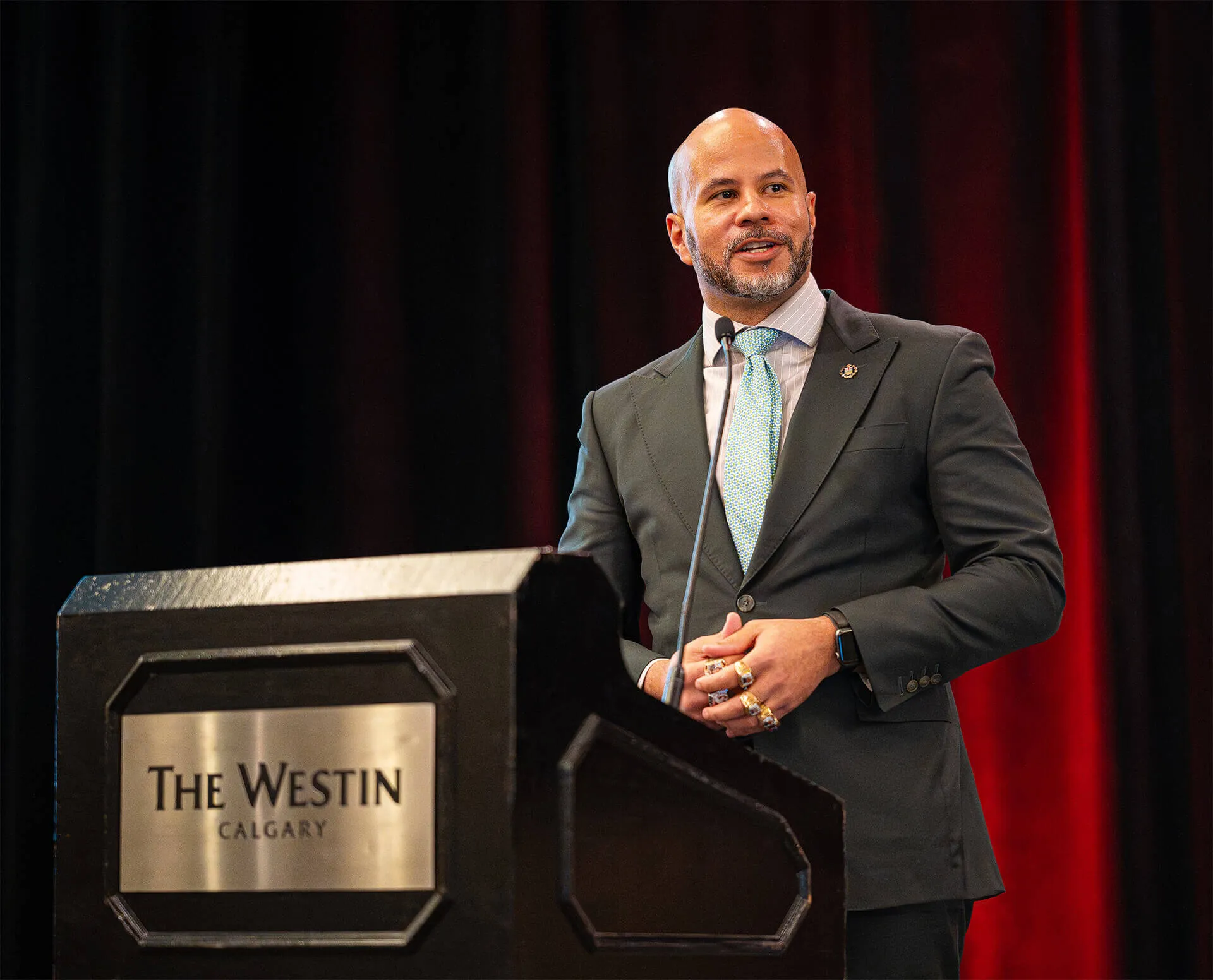 Man in a suit speaking at a podium with The Westin Calgary plaque, with a black and red curtain backdrop.