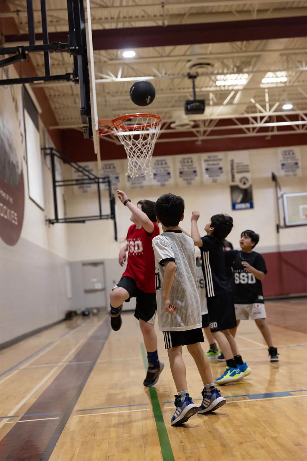 Young boys playing basketball in a gymnasium with one boy in a red jersey jumping to shoot the ball towards the hoop.