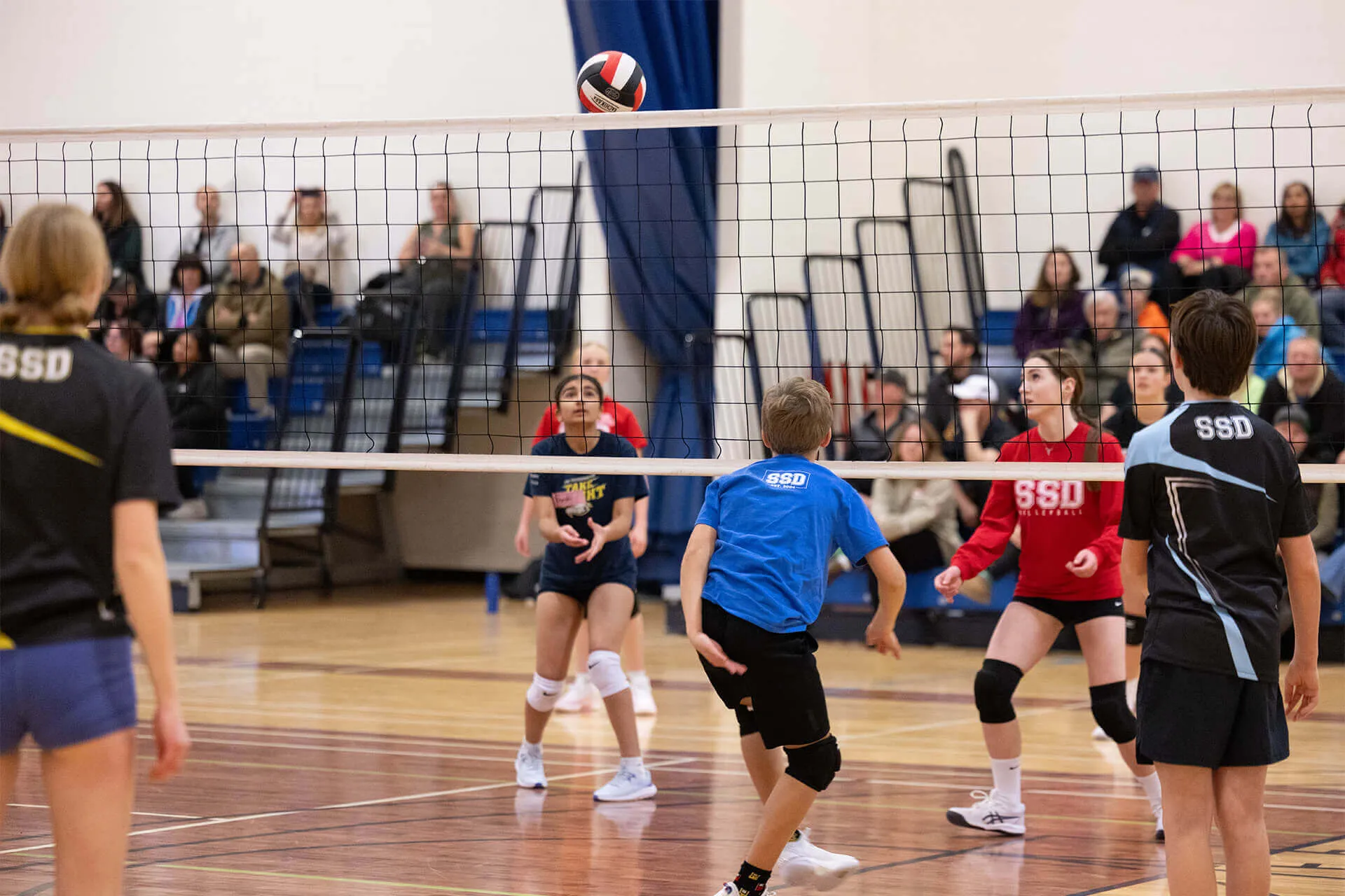 Youth volleyball players focused on a ball mid-air above the net during an indoor game with spectators in the background.