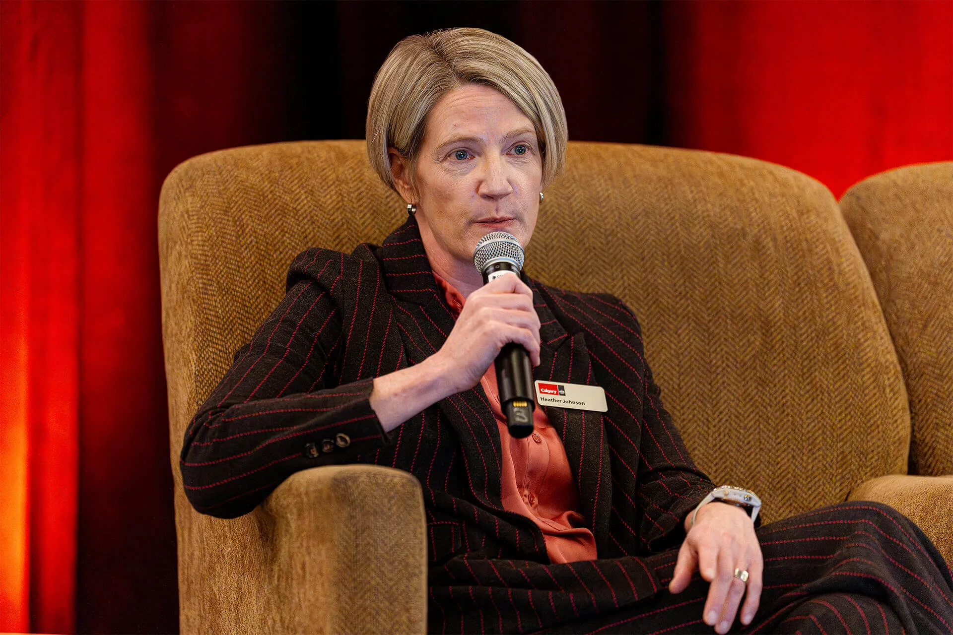 Woman with short blonde hair wearing a dark pinstripe suit and a name tag, seated in a brown chair, speaking into a microphone.