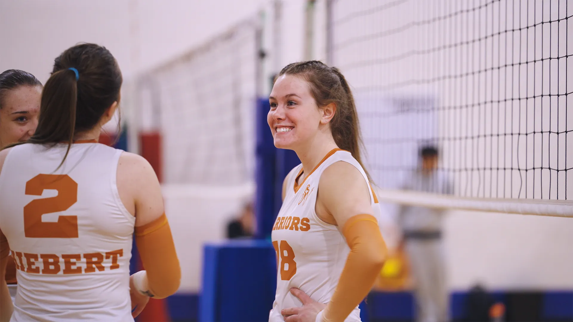 Smiling female volleyball player wearing a white and orange jersey with number 18 and the word Warriors, standing with hands on hips near the net, talking to teammate wearing number 2 jersey.