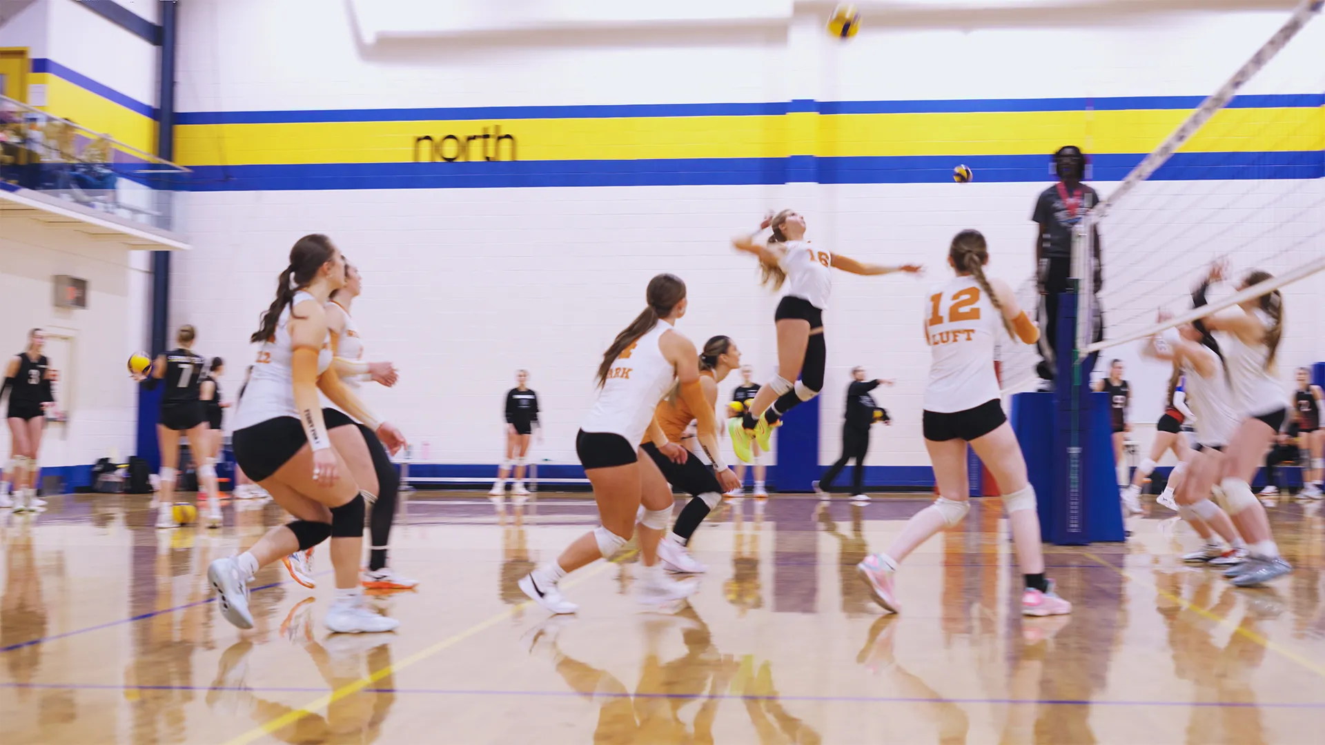 Indoor volleyball game with female players in white and black uniforms, one player jumping to hit the ball over the net, others watching or preparing to react.