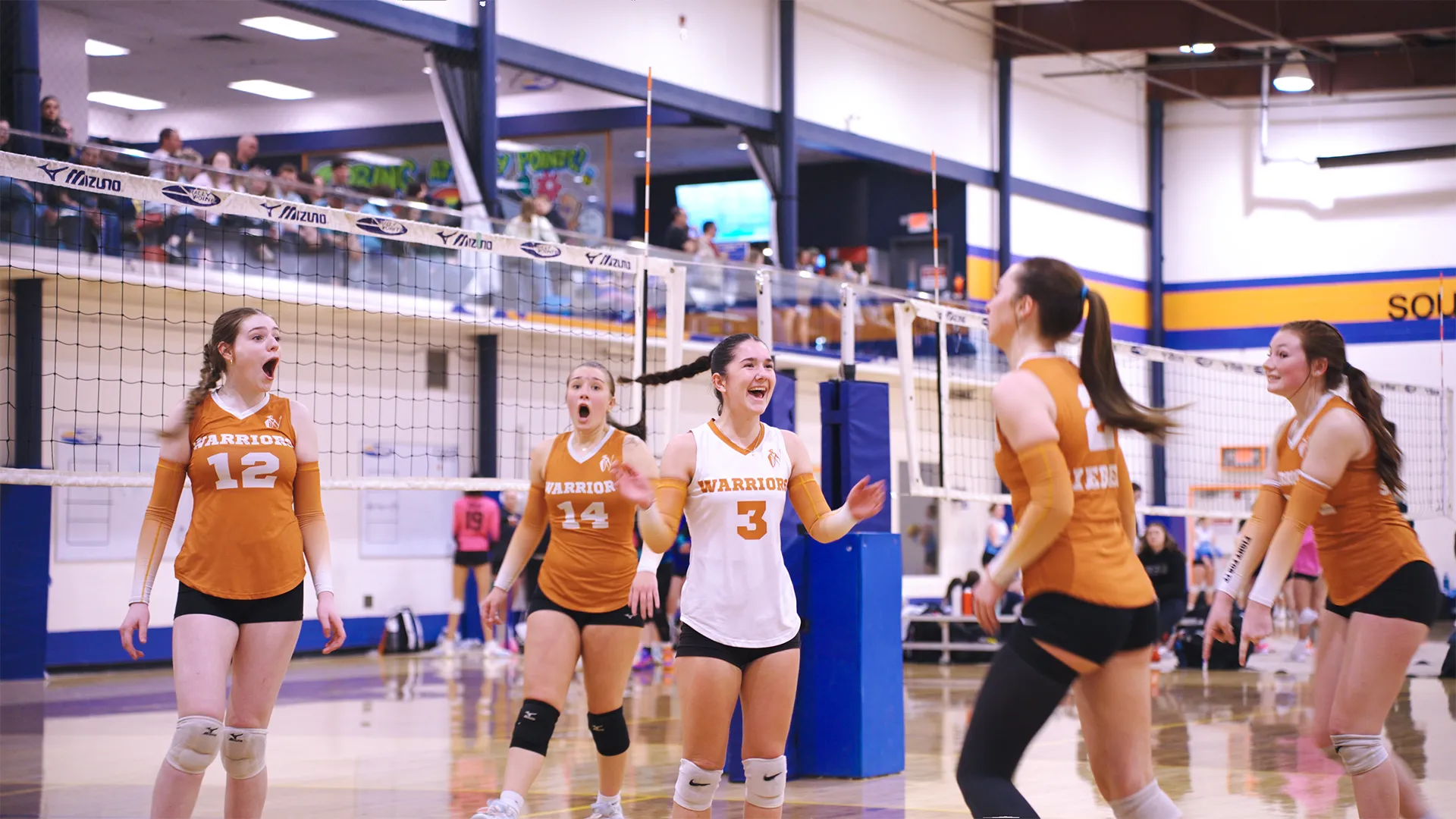 Five female volleyball players on a court wearing orange and white uniforms marked 'Warriors' react with surprise and joy near the net during a game in a gymnasium with spectators in the background.