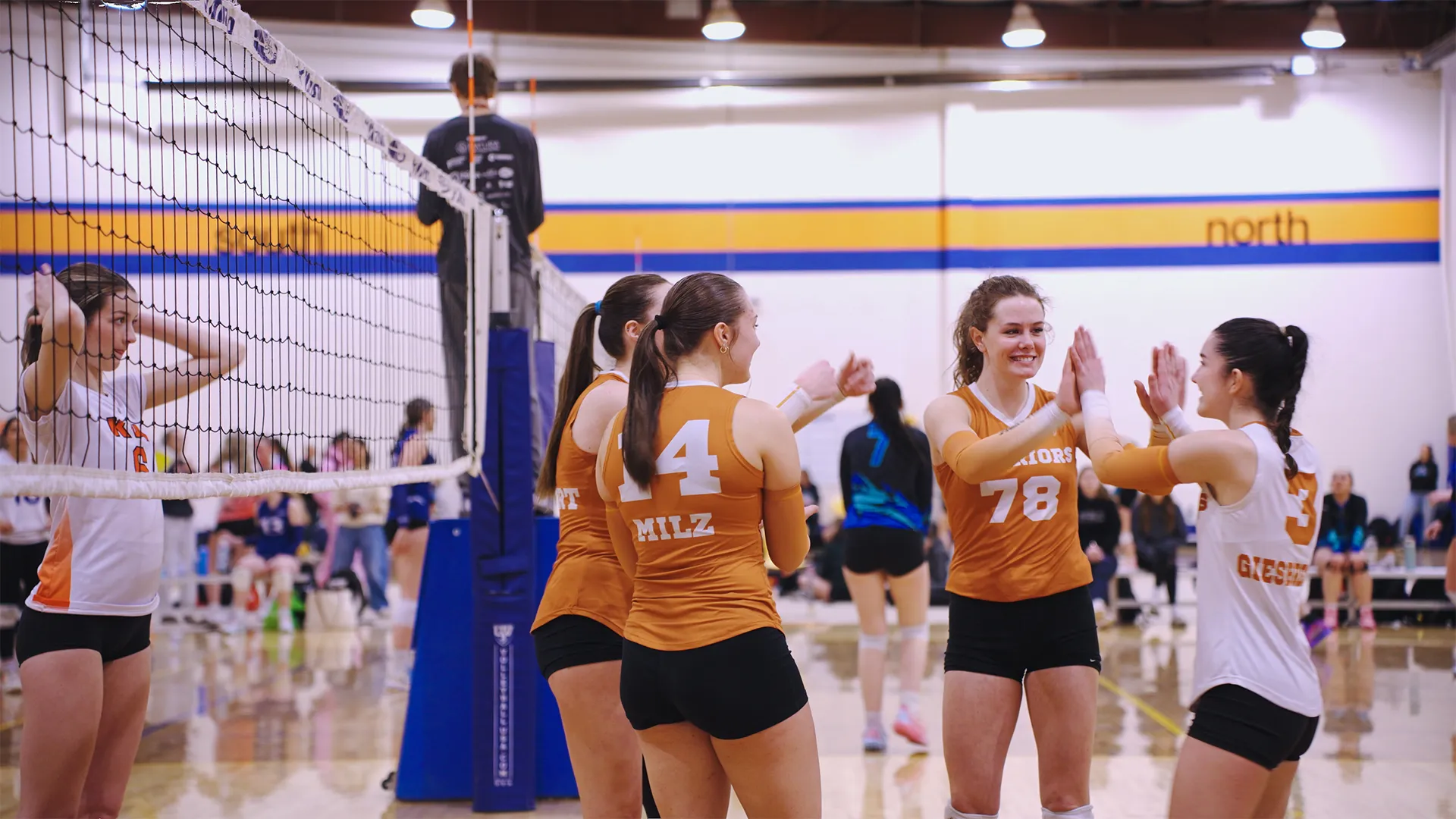 Female volleyball players on a court, some in orange jerseys celebrating with high fives, others standing near the net and waiting, with a referee visible in the background.