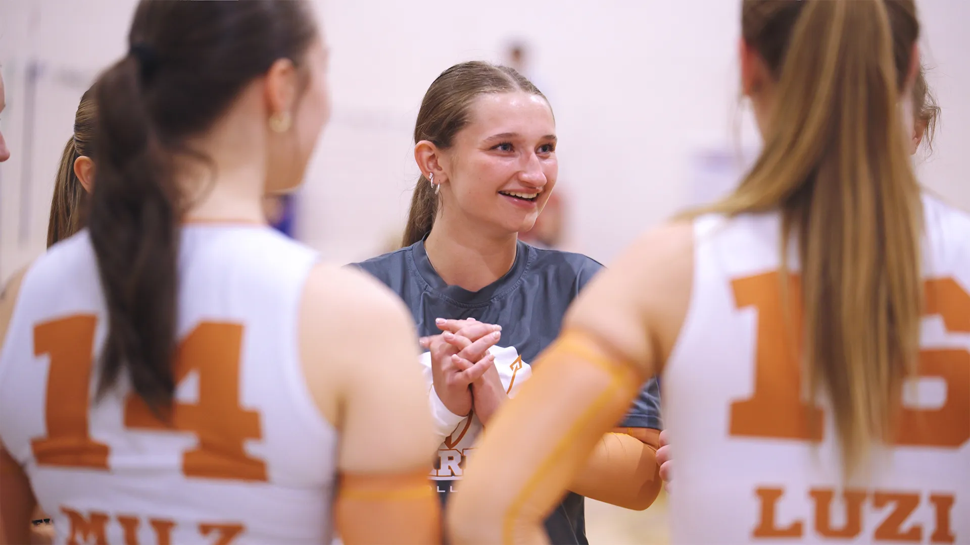 A young female volleyball player with a ponytail and hoop earrings smiles and clasps her hands while talking to teammates wearing white jerseys numbered 14 and 16.