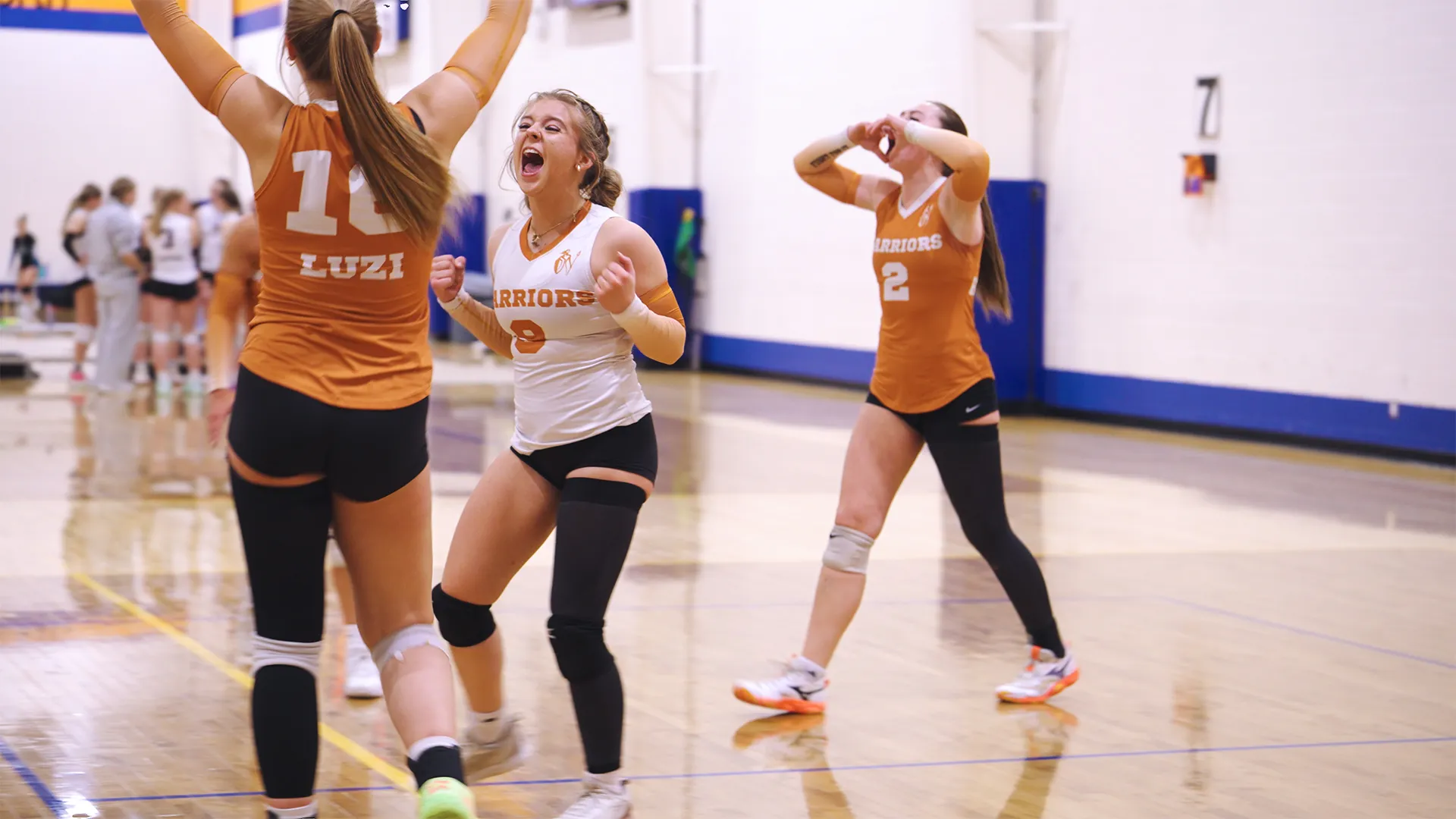 Three female volleyball players celebrating on an indoor court with one player in a white jersey and the other two in orange jerseys, showing excitement and joy.