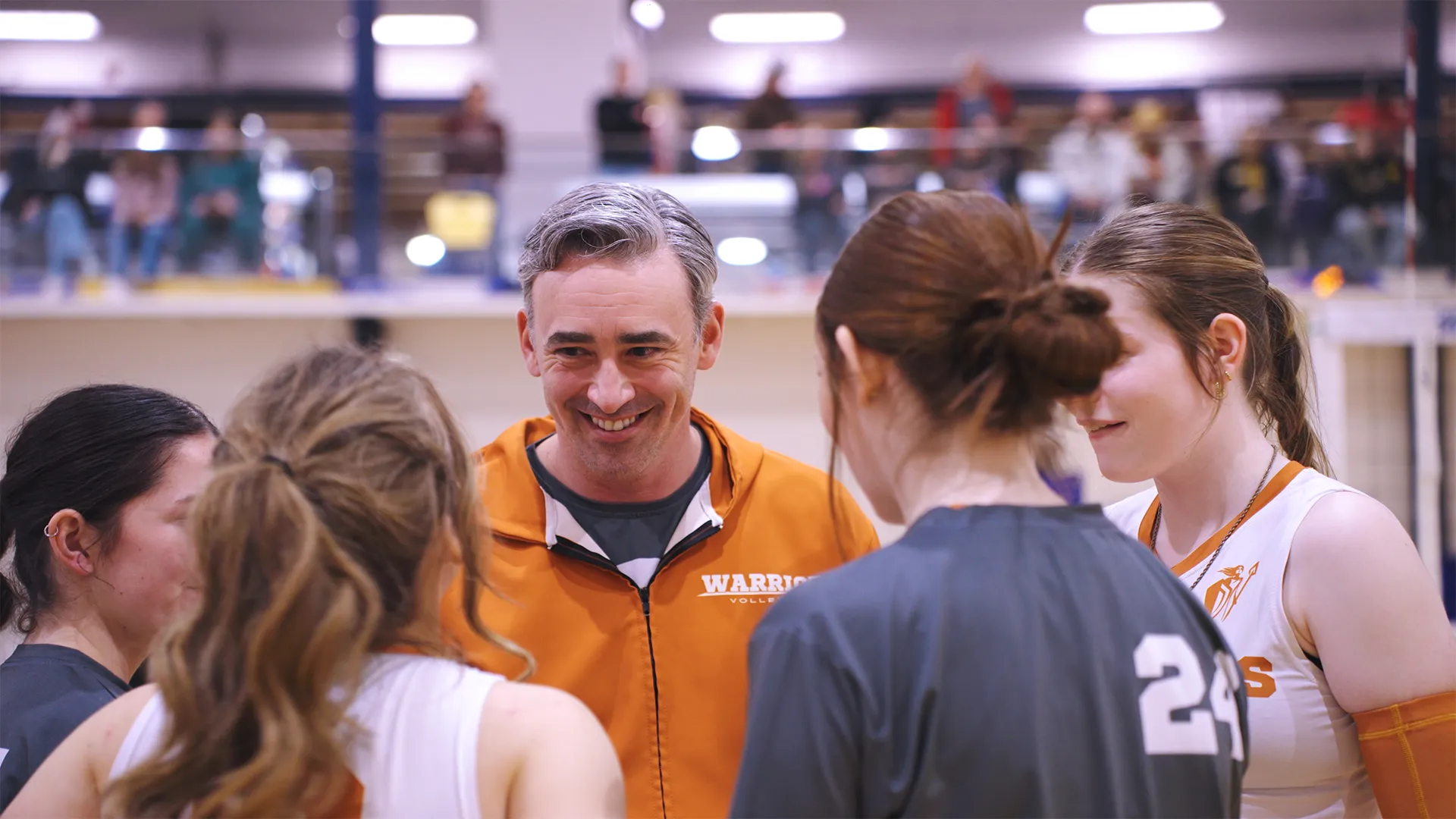 A male volleyball coach in an orange jacket speaking and smiling to four female volleyball players in a gym with spectators in the background.
