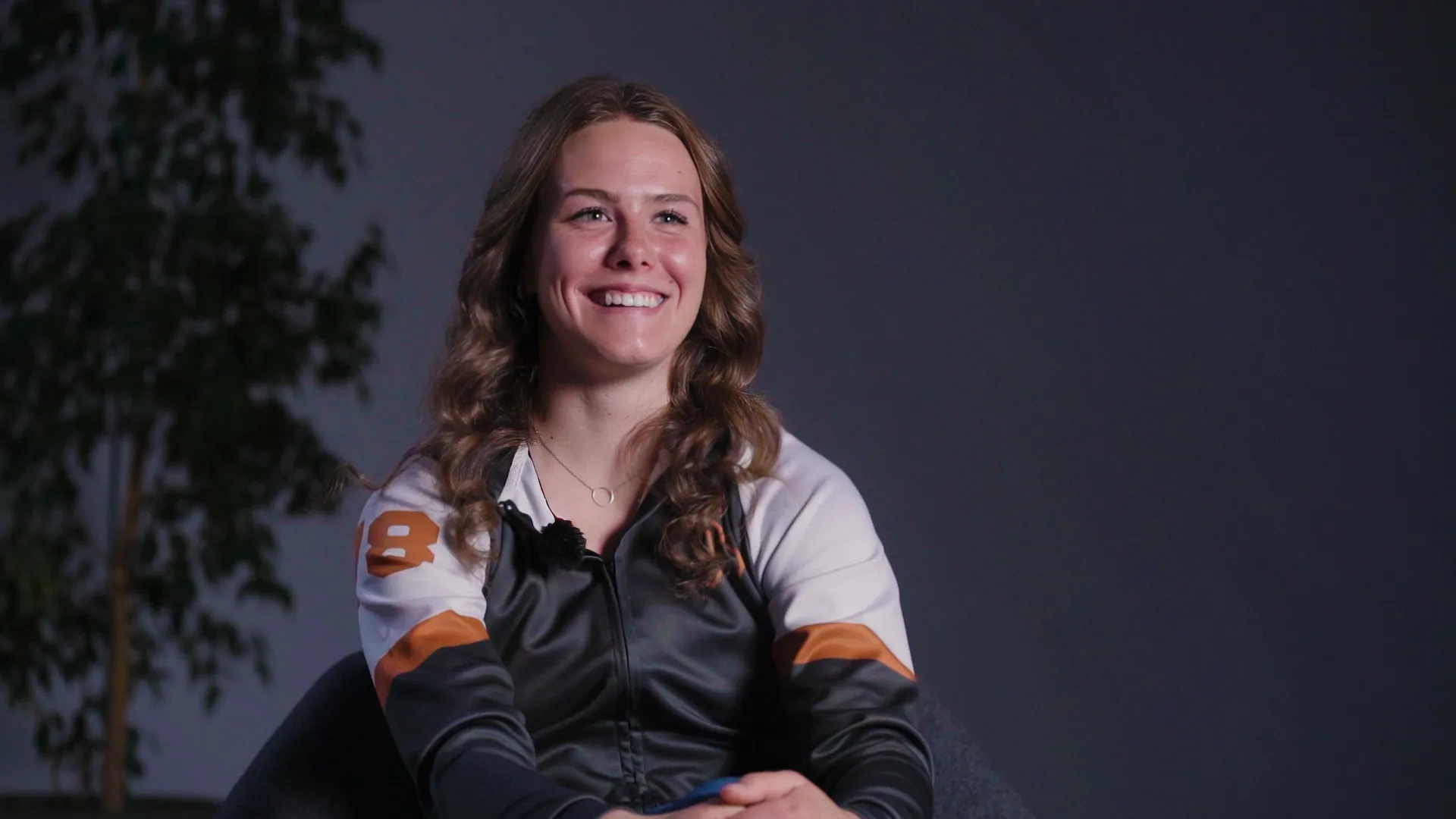 Smiling young woman with wavy brown hair wearing a black, white, and orange jacket, seated against a dark background with a plant partially visible on the left.