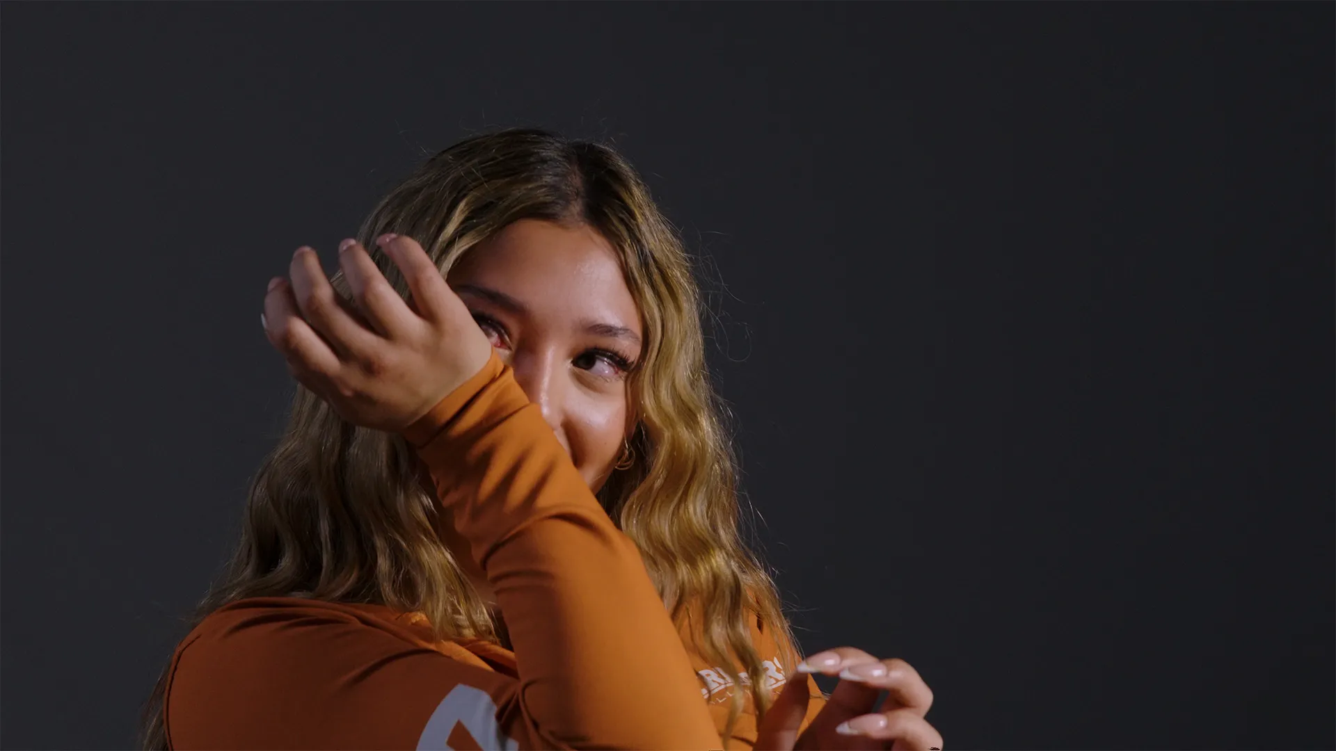Young woman with long wavy blonde hair wearing an orange top wiping tears from her eyes with her arm against a dark background.