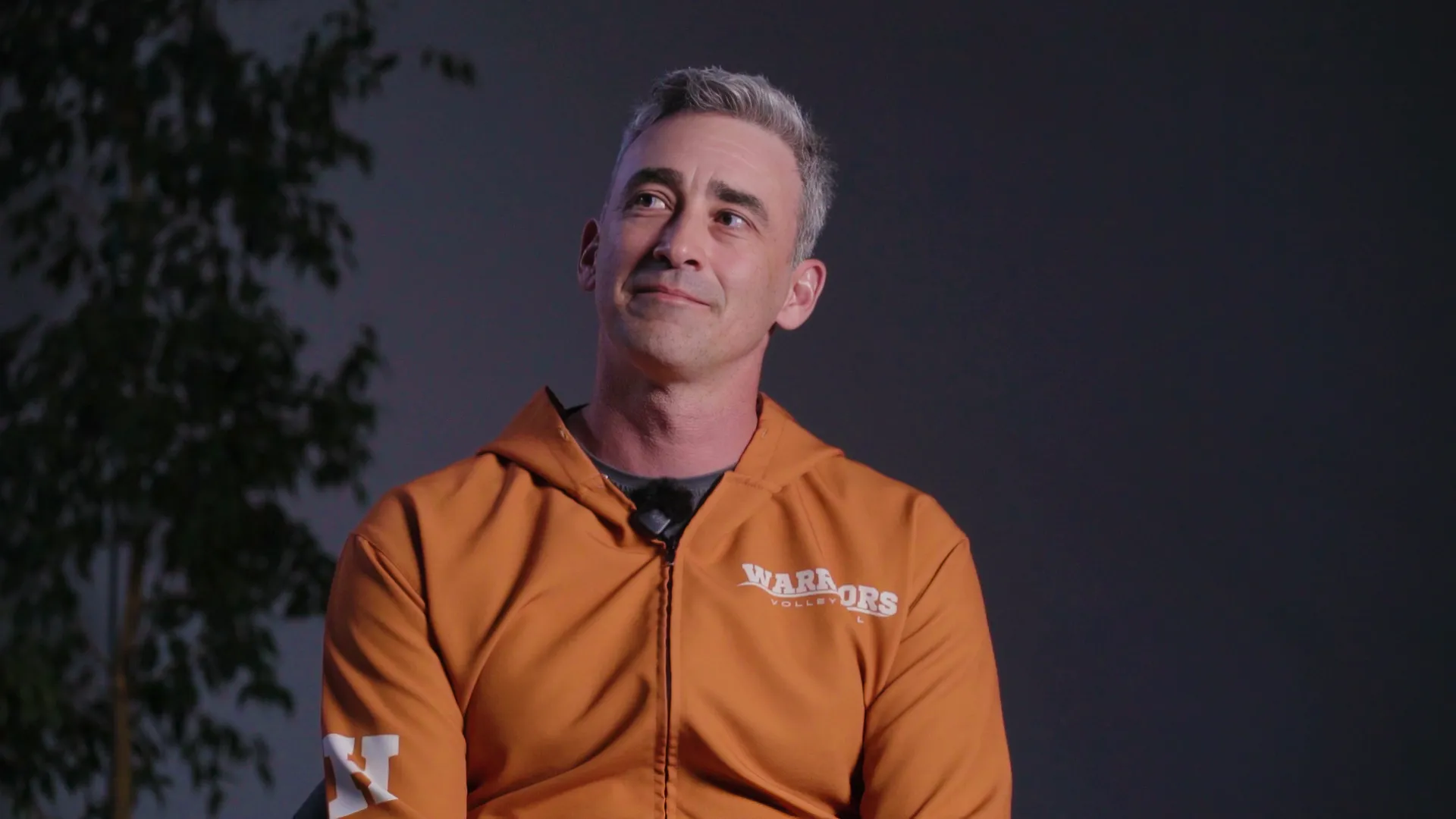 Man with short gray hair wearing an orange jacket with 'WARRIORS VOLLEYBALL' text, looking slightly upward with a subtle smile against a dark background with a blurred plant.