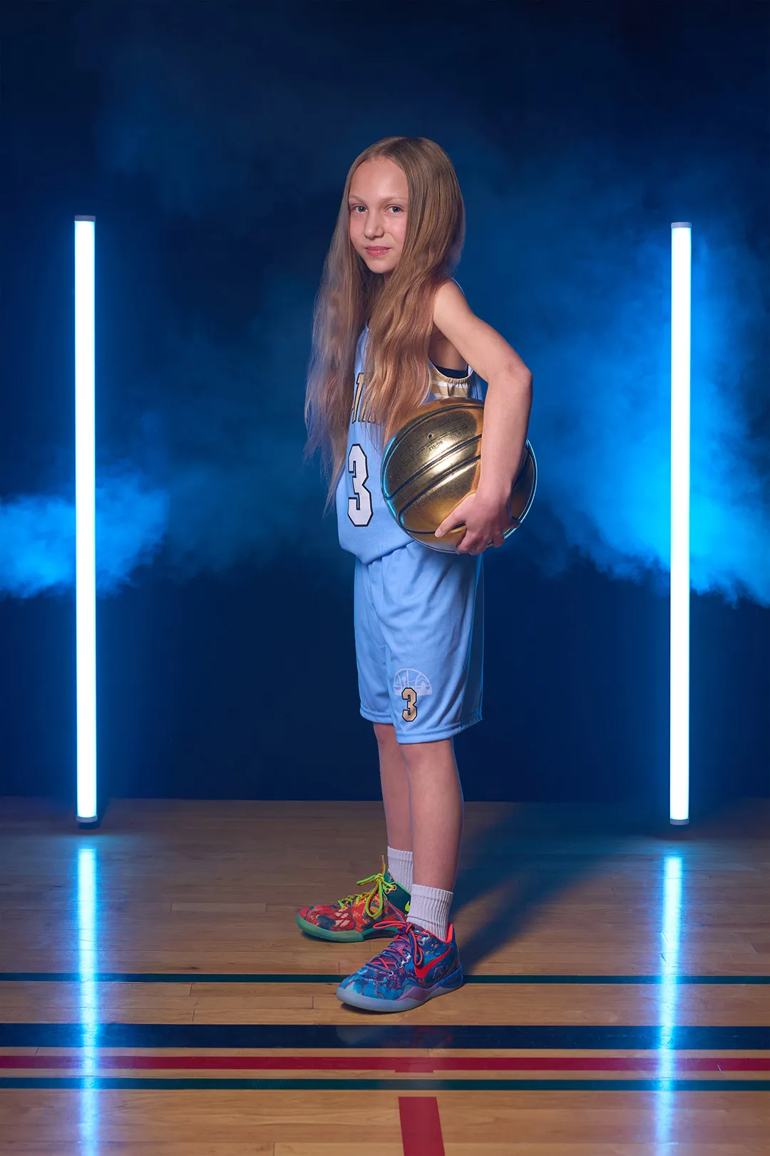Young girl with long blonde hair in a blue basketball uniform holding a gold basketball under one arm, standing on a gym floor with colored lines, flanked by two vertical blue neon lights and blue fog in the background.