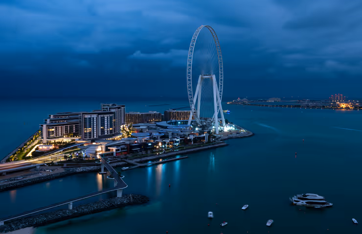 Aerial night view of Bluewaters Island in Dubai, featuring the Ain Dubai observation wheel lit up against the dark blue waters and evening sky.