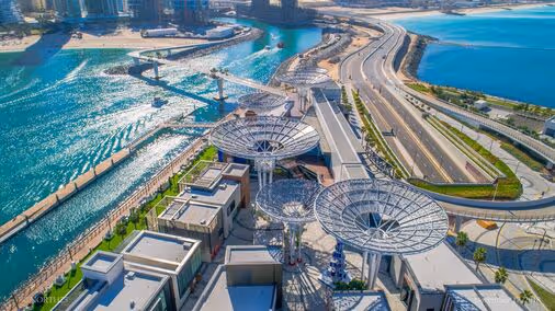Aerial view of a futuristic coastal development with modern architecture, wide roads, and turquoise waters, likely part of Dubai Marina or Bluewaters Island.