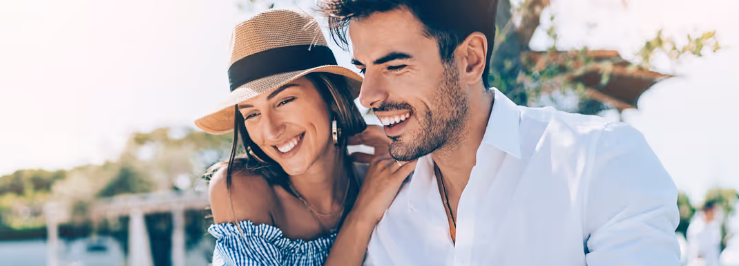 Smiling couple enjoying a sunny day outdoors, dressed in light summer clothing, with trees and bright sky in the background.