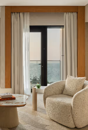 Cozy modern lounge area with a plush armchair and small table, featuring a window view of the sea beyond a balcony.