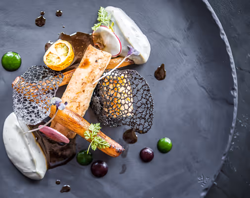 Gourmet fine dining dish artfully plated with creative textures, garnishes, and sauces on a dark plate.