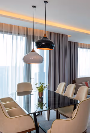 Modern dining area with a glass-top table, cushioned chairs, pendant lights, and sheer curtains letting in natural light.