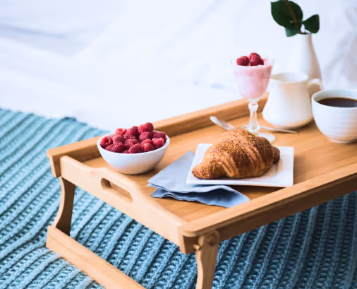 Breakfast in bed setup with a wooden tray holding a croissant on a plate, a bowl of raspberries, a glass of yogurt with berries, a teapot, and a cup of coffee on a blue textured blanket.