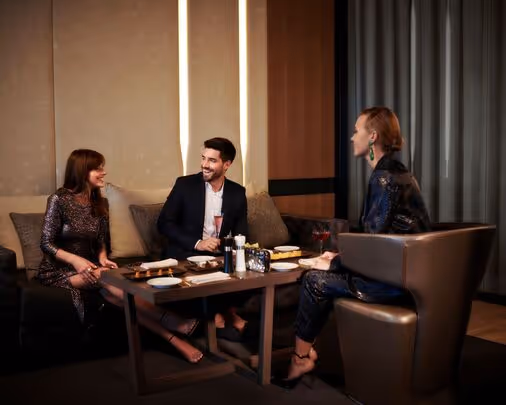 Three people sit in a modern office or lounge, engaged in conversation. Two sit on a couch facing a woman who sits across from them in an armchair. A coffee table with documents is between them.