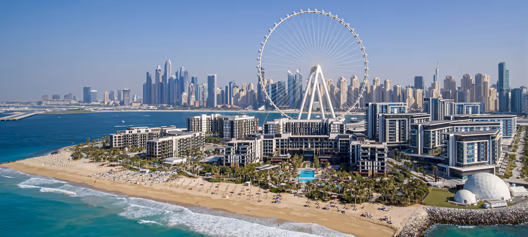 Aerial view of a sandy beach with rows of umbrellas, a luxury resort, palm trees, and a large Ferris wheel, set against the Dubai city skyline and blue coastline.