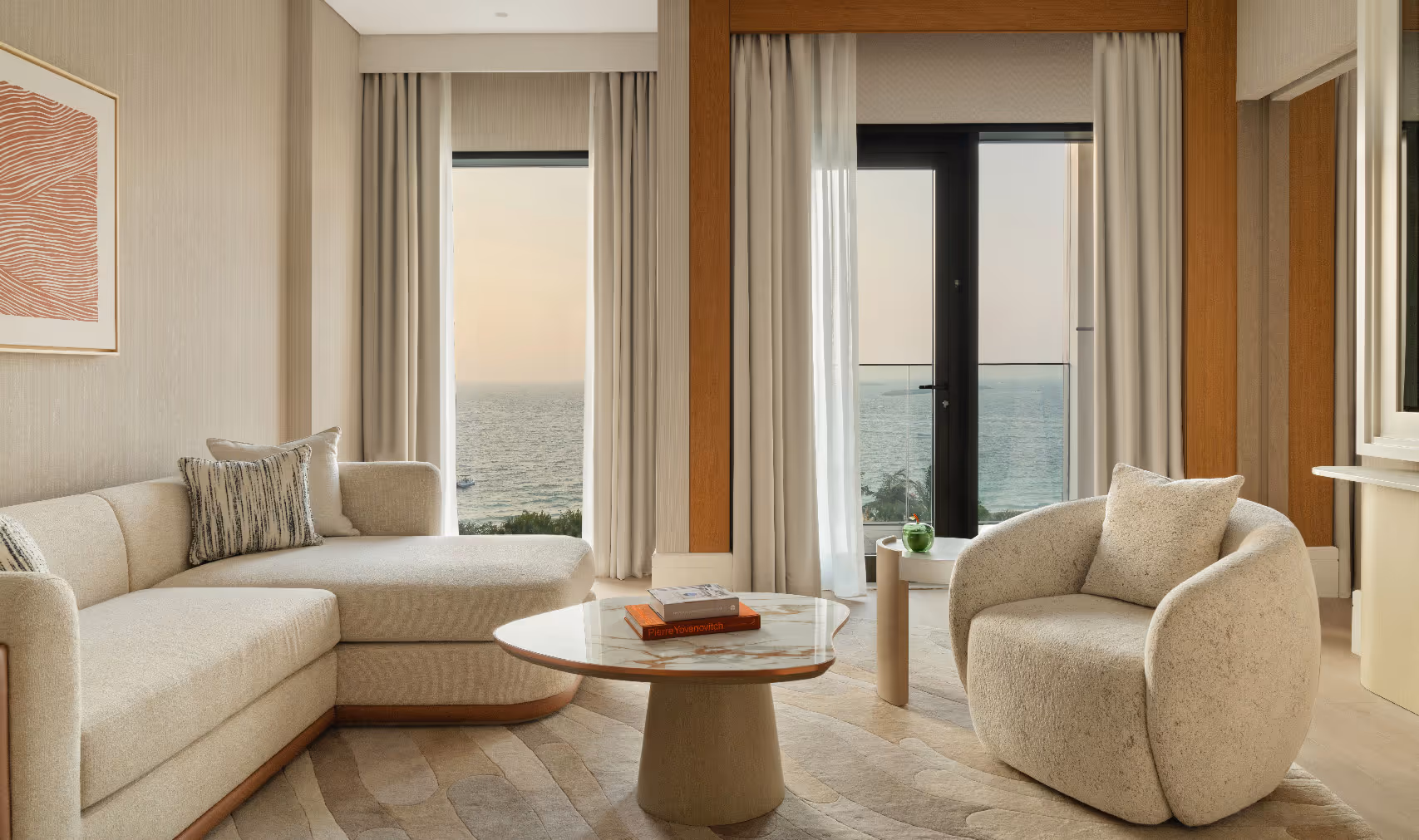 A modern, neutral-toned living room with a curved sofa, plush armchair, round marble coffee table, and large windows offering an ocean view through light curtains at sunset.


