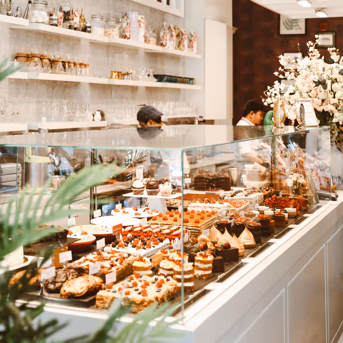 A bakery display case filled with assorted pastries, cakes, and desserts, with two people working behind the counter. The shop has white shelves, glass jars, decorative plants, and a large floral arrangement.