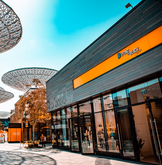 A modern outdoor shopping area with sleek glass storefronts, orange accents, geometric metal canopies, and people walking or sitting near shops under a bright turquoise sky.