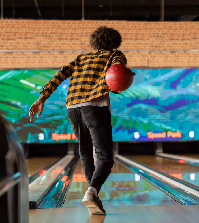 A person wearing a yellow and black checkered shirt bowls a red ball down a lane at an indoor bowling alley with colorful lights in the background.