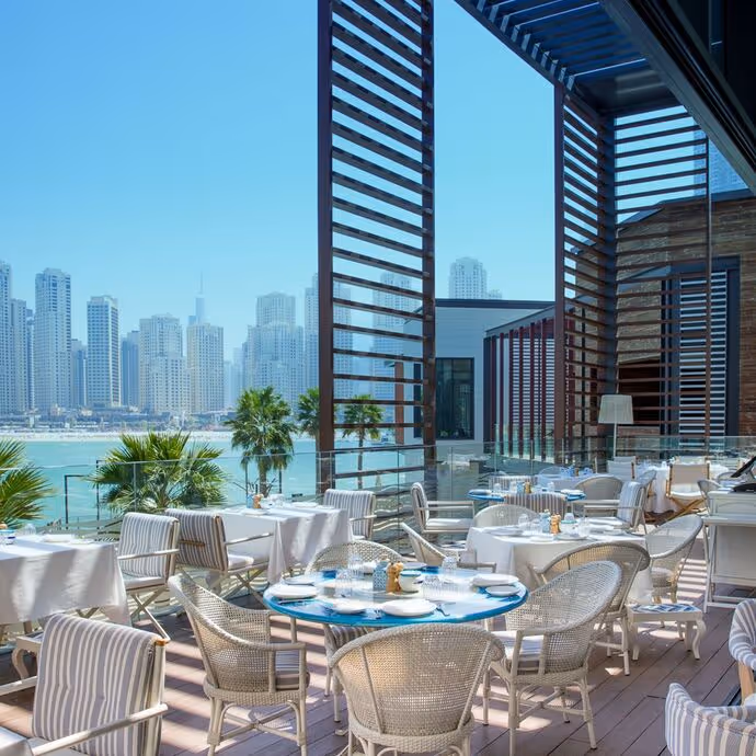 Outdoor restaurant with white tables and wicker chairs overlooks a waterfront with palm trees and a skyline of modern high-rise buildings under a clear blue sky.



