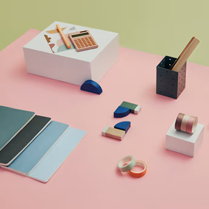 Minimalist arrangement of stationery items including notebooks, tape, swatches, and a pencil holder on pastel pink and green background.