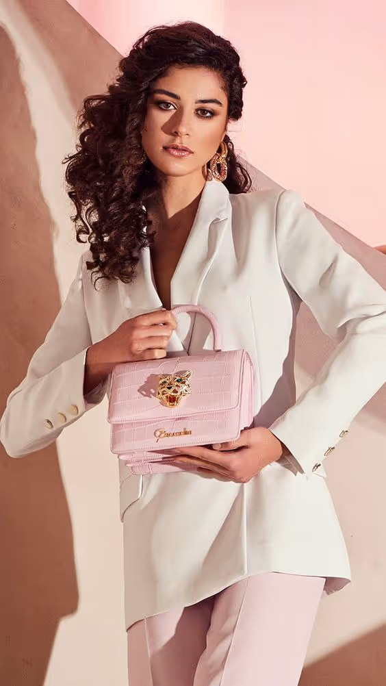 A woman with curly dark hair wears a white blazer and pale pink pants, holding a light pink handbag with a gold embellishment, standing against a soft pink and beige background.