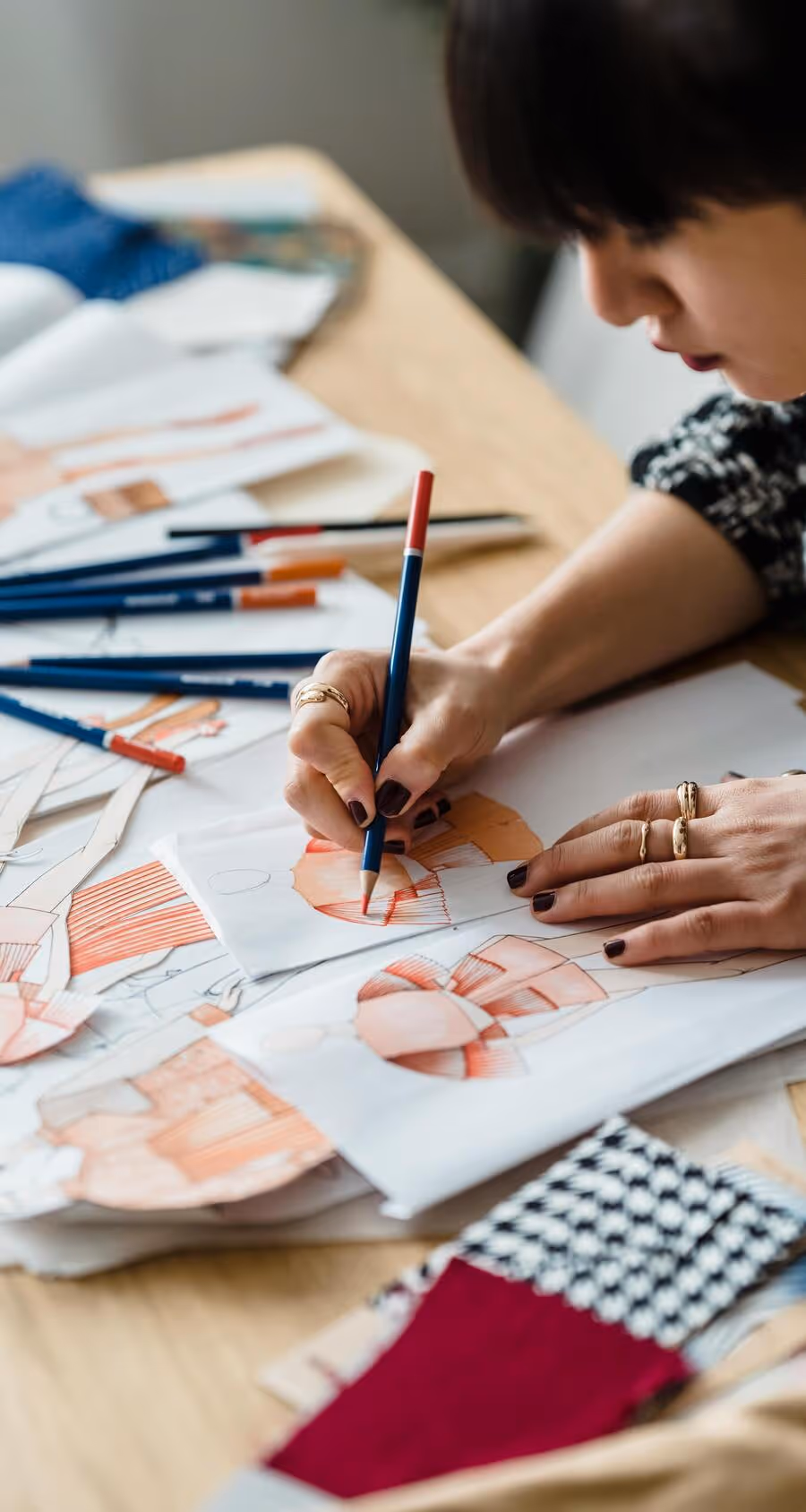 Person sketching fashion designs on paper surrounded by colored pencils, fabric swatches, and design materials on a desk.