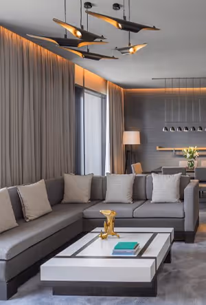 Modern living room with a large gray sectional sofa, beige pillows, and a white coffee table with decorative gold accents, illuminated by contemporary ceiling lighting

