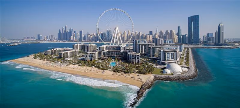 Aerial view of a modern beachfront development with a large Ferris wheel and a city skyline in the background.